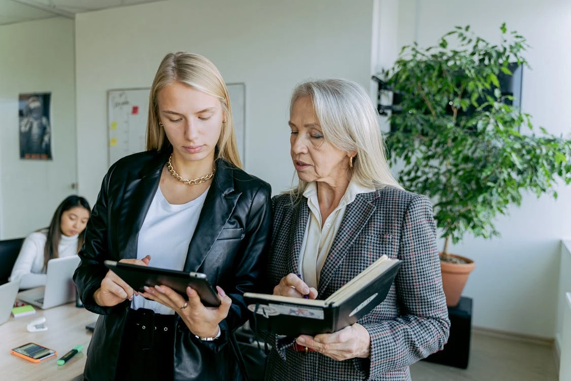 Two women in business attire, one holding a tablet and the other holding a notebook, engaged in a discussion in an office setting.