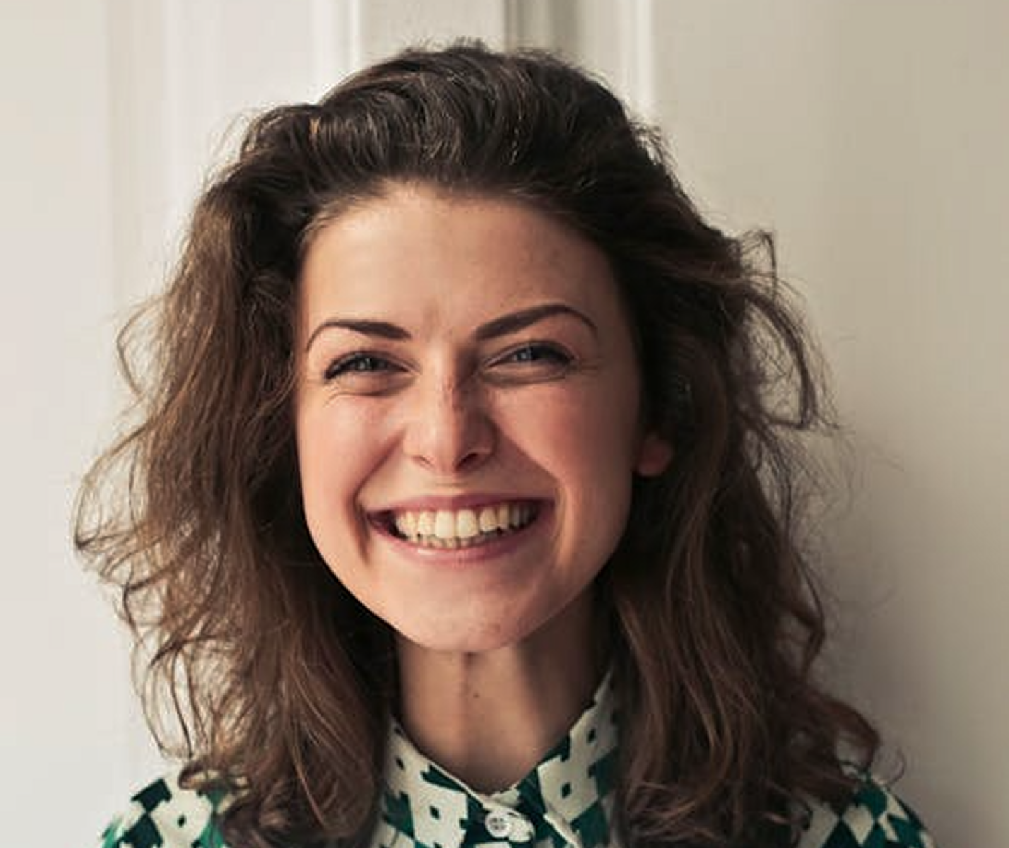 Close-up of a smiling woman with curly brown hair wearing a patterned collared shirt.