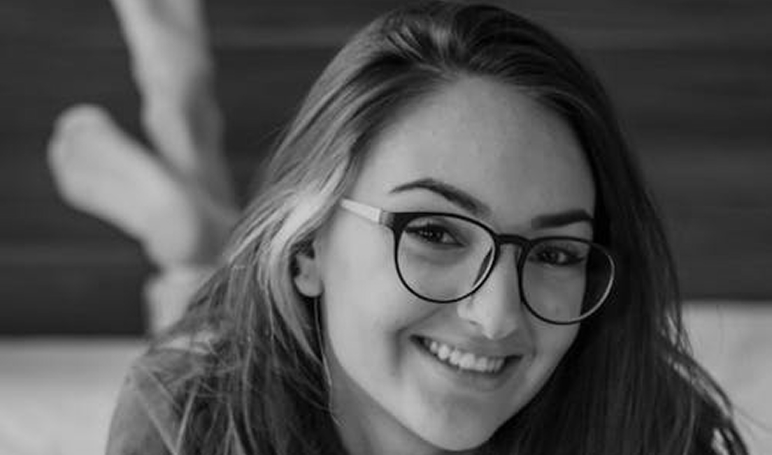 Close-up of a smiling young woman wearing large black-framed glasses lying down with feet raised behind her.