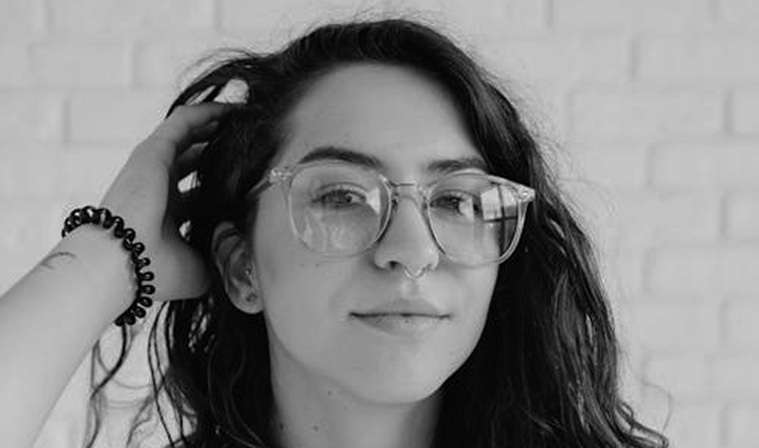 Close-up of a woman with long dark hair, wearing large transparent glasses and a nose ring, with a hand running through her hair.