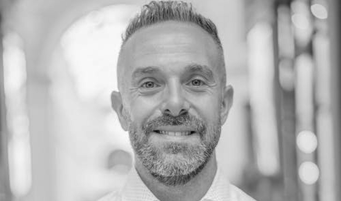 Smiling man with short hair and beard, wearing a collared shirt in a softly blurred indoor setting.