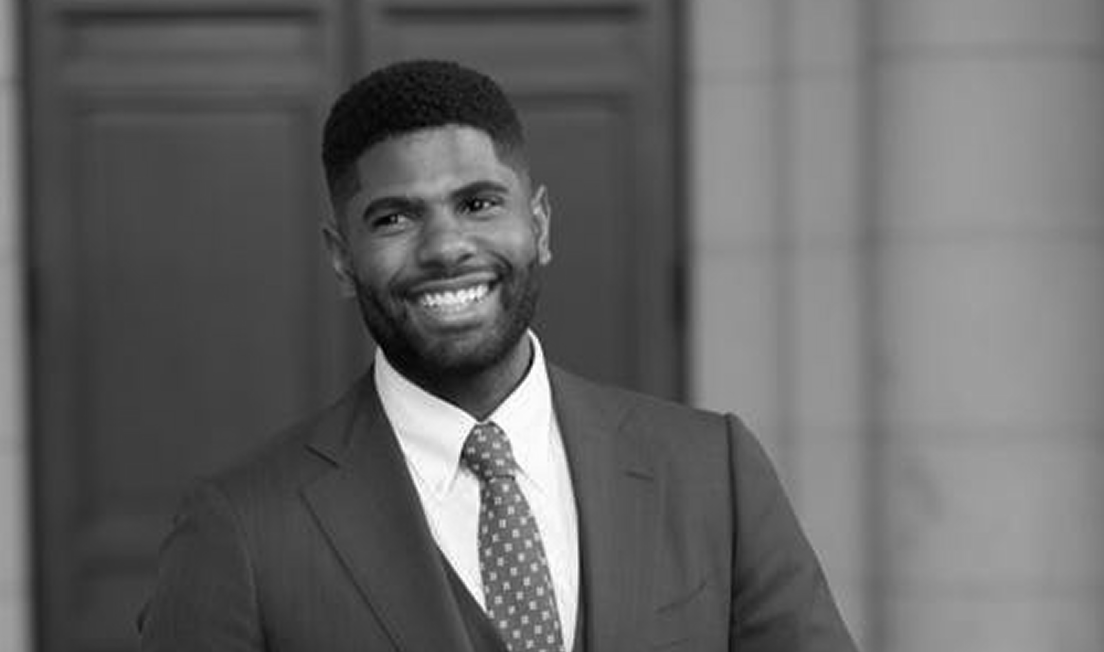 Smiling man with short curly hair and beard wearing a suit and patterned tie standing in front of a building entrance.