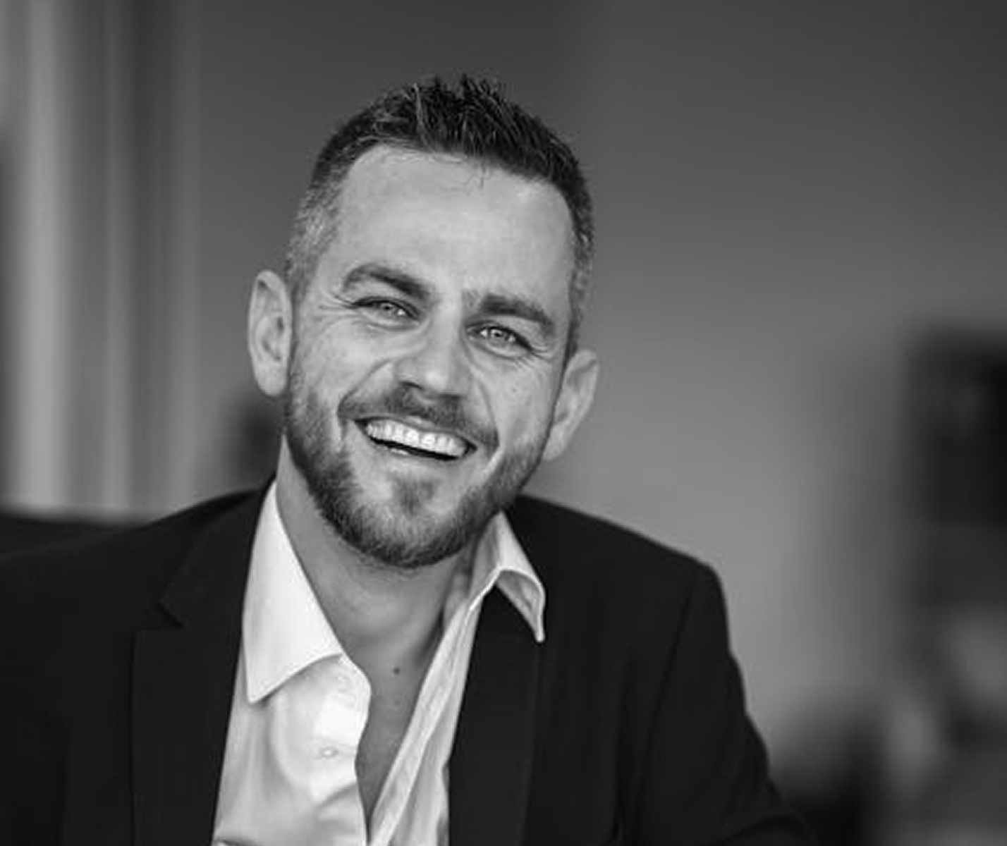 Smiling man with short hair and beard wearing a white shirt and dark blazer, black and white photo.
