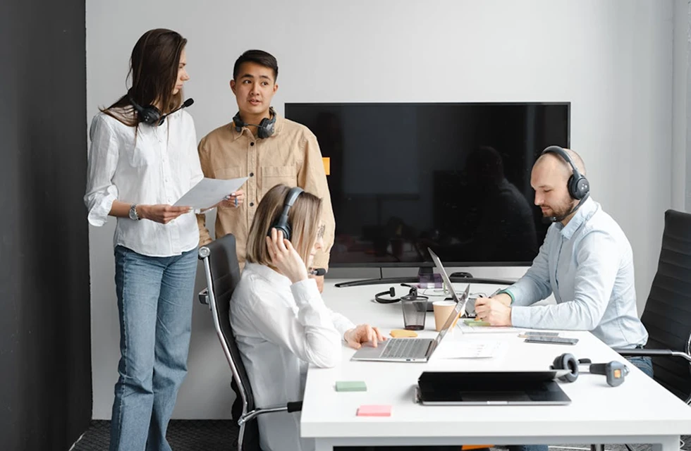 Two women standing indoors, one holding a tablet and the other a planner, engaged in discussion.